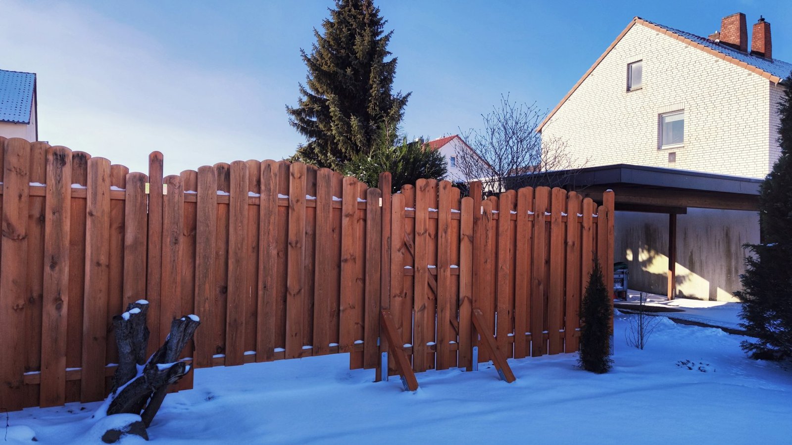 Red-stained picket fence around a snow-covered back garden, a house behind it